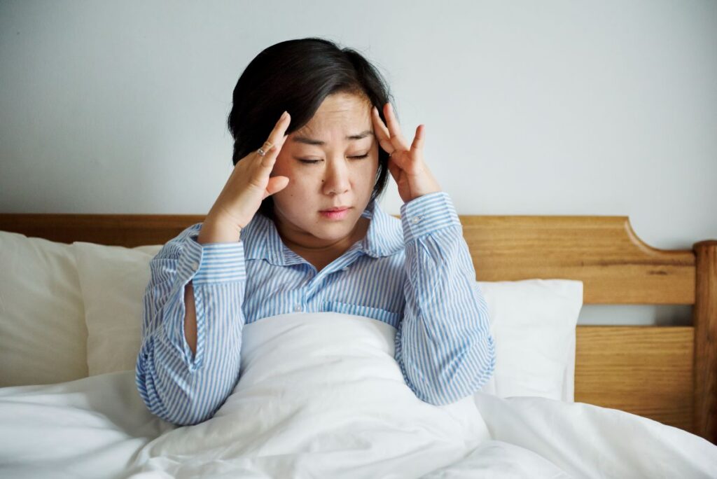 Woman sitting up in bed with white blankets, rubbing her temples as if in pain or stress.