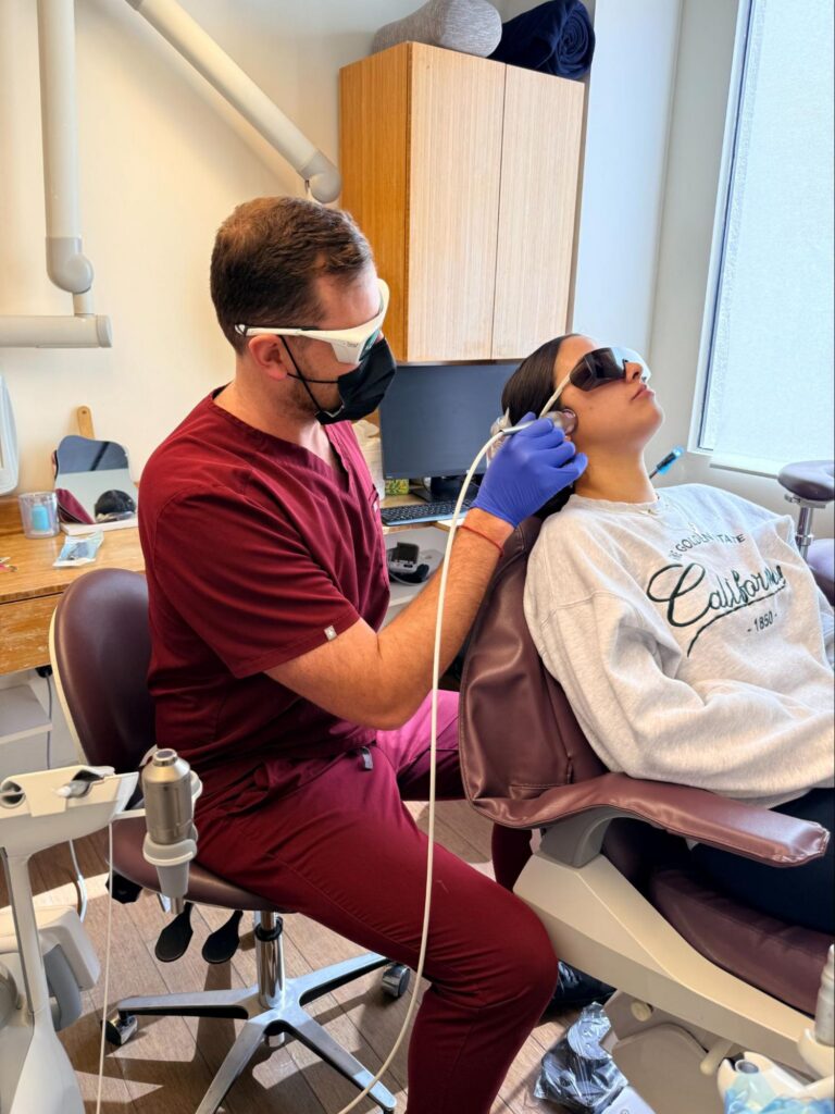 Dentist in maroon scrubs and mask uses dental tool on patient in reclining chair in a clinic setting with cabinets above.