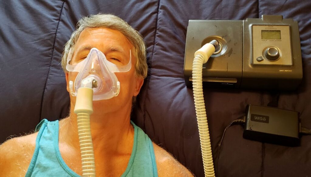 Man lying in bed wearing a CPAP mask connected to a sleep apnea machine on a dark blue blanket/pillow.