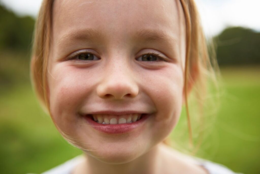 Close-up of smiling young girl with light brown hair outdoors in a green field.