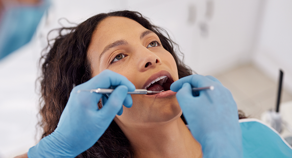 Patient receiving dental care: close-up of a woman with mouth open as blue-gloved hands hold dental instruments inside her mouth in a dental chair setting.