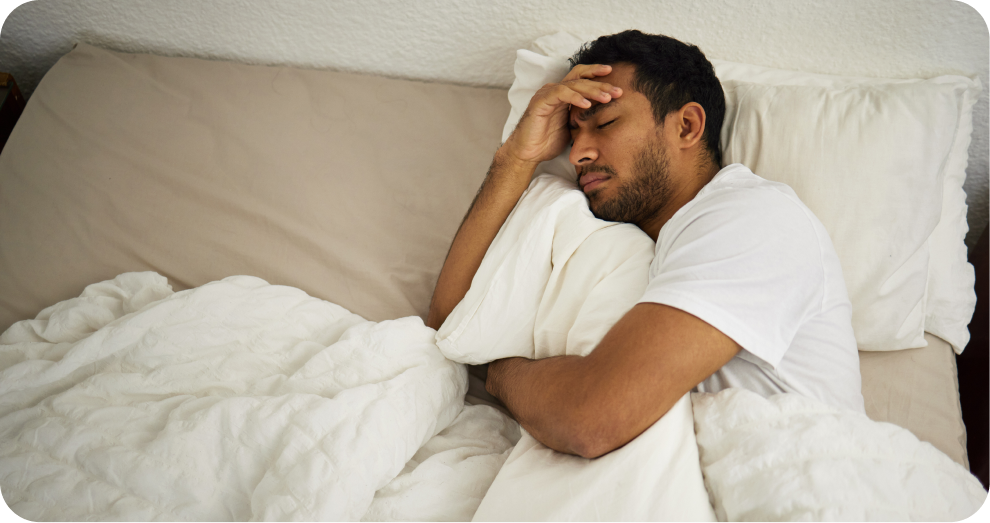 Man lying in bed on his side with a hand on his forehead, looking unwell or tired under a white blanket and pillows.